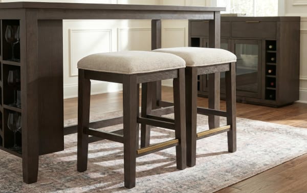 Dark wood bar stools with beige cushioned seats and brass footrests under a matching dark wood bar table on a patterned rug.