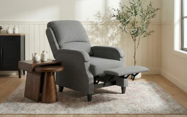 Gray fabric recliner chair with padded armrests and extended footrest, paired with dark wood nesting tables, set on a patterned area rug in a sunlit living room.