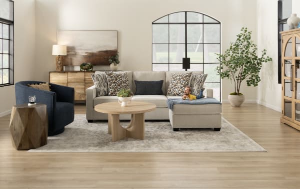 Modern living room with beige sectional sofa, patterned cushions, wooden coffee table, blue armchair, potted plant, and wall art.