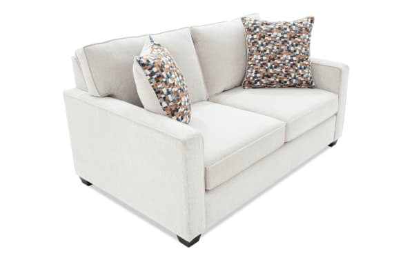 Light beige fabric loveseat sofa with two patterned throw pillows, featuring cushioned seats and backrests, modern design with clean lines.