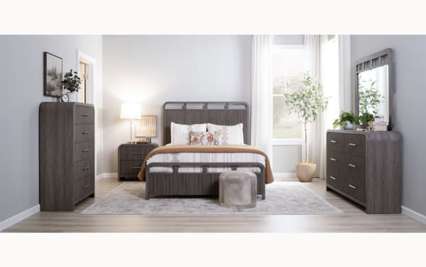 Modern bedroom set with gray wood finish including bed frame with headboard and footboard, matching dresser, chest of drawers, nightstand, and mirror. Light wood flooring, neutral area rug, beige bedding with decorative pillows, and potted plants by window create a cozy, contemporary style.