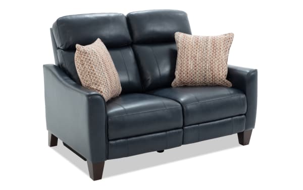 Black leather reclining loveseat with two patterned beige throw pillows, featuring padded armrests and wooden tapered legs, ideal for modern living rooms.