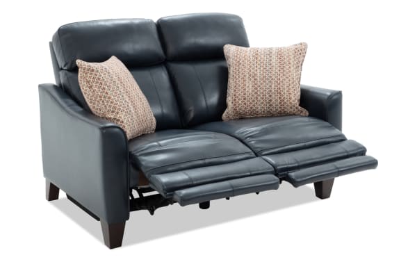 Black leather reclining loveseat with two patterned beige throw pillows, padded armrests, and wooden legs, ideal for modern living rooms.