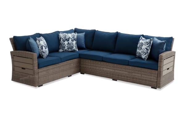 Sectional outdoor patio sofa with brown wicker frame and navy blue cushions, featuring decorative leaf-patterned throw pillows.