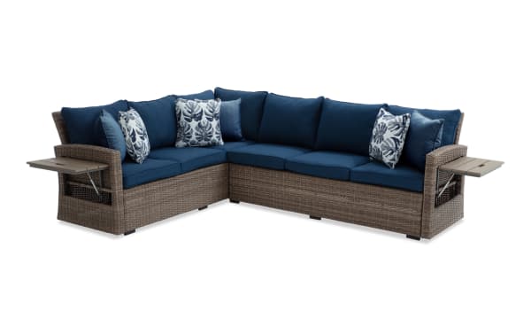 Outdoor sectional sofa with brown wicker frame, navy blue cushions, and leaf-patterned throw pillows, featuring foldable side tables.