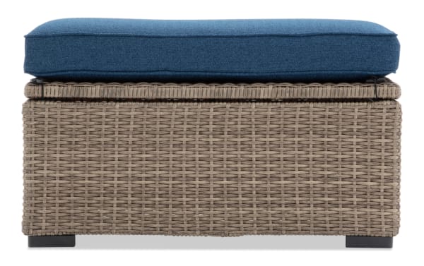 Outdoor wicker storage bench with a woven rattan design, featuring a blue cushioned seat and black feet for patio or garden use.