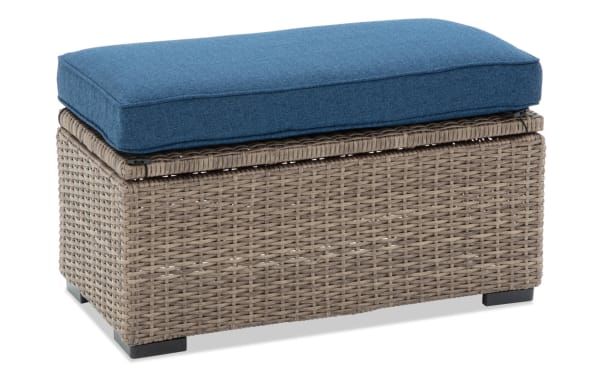 Wicker storage bench with a blue cushioned top, featuring woven rattan design and black feet, suitable for indoor or outdoor seating and storage.