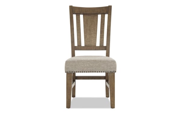 Wooden dining chair with beige upholstered seat, nailhead trim, and slatted backrest, featuring a rustic finish and sturdy legs.