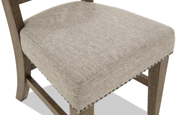 Upholstered dining chair seat with beige textured fabric and nailhead trim, supported by rustic wooden legs with crossbars.