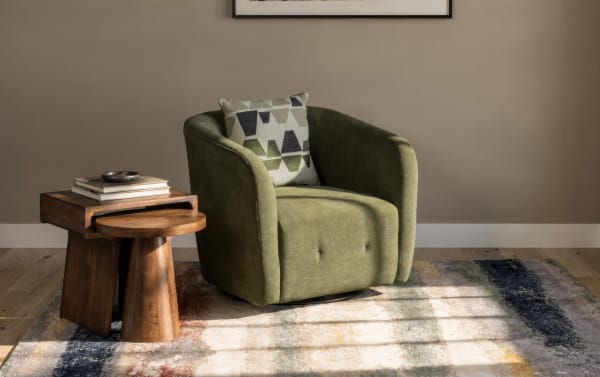 Green upholstered swivel armchair with a geometric patterned cushion, paired with nested wooden side tables on a multicolor area rug in a minimalist living room.