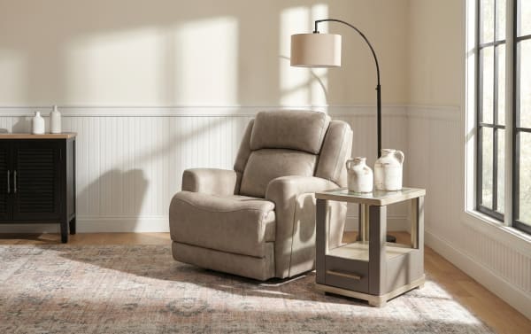 Beige leather recliner chair with plush cushioning, paired with a modern beige side table with glass top and ceramic pitchers, set in a sunlit living room with wood flooring and a patterned area rug.