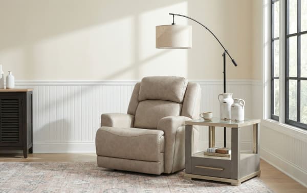 Beige leather recliner chair with padded armrests and headrest, paired with a modern gray and beige side table featuring a drawer and glass top, next to a black arc floor lamp with a beige fabric shade, set in a bright room with large windows and light wood flooring.