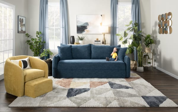 Modern living room with blue fabric sofa, yellow upholstered armchair, green potted plants, abstract wall art, and geometric patterned rug.