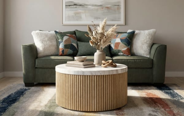 Green velvet sofa with patterned and fluffy white throw pillows, a round wooden coffee table with a marble top, decorative vase, and books in a modern living room.