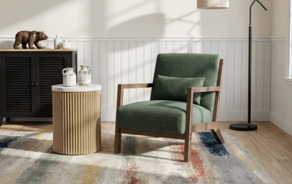 Mid-century modern green upholstered armchair with wooden frame and matching lumbar pillow, next to a round wooden side table with ceramic jugs, on a multicolor patterned rug in a bright living room.