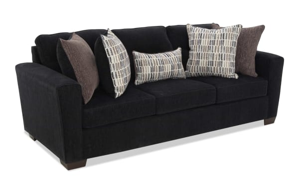Black fabric sofa with three seat cushions and wide armrests, featuring six decorative pillows in brown and patterned beige tones, supported by short wooden legs.