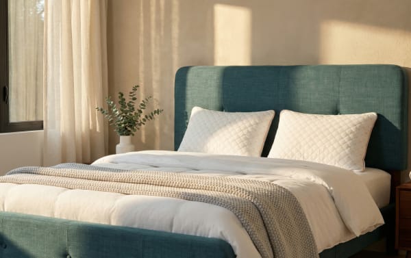 King-size bed with teal upholstered headboard and footboard, white quilted pillows, white bedding, and beige knitted throw in sunlit bedroom.