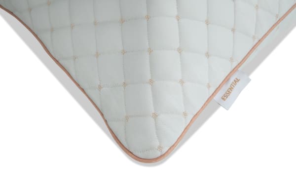 Corner of a quilted white pillow with beige stitching and beige piping along the edge, featuring a white tag with the word "ESSENTIAL" in beige text.