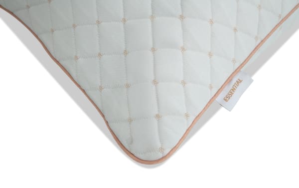 Corner of a quilted white pillow with beige stitching and beige piping, featuring a fabric tag with the word "ESSENTIAL" in beige text.