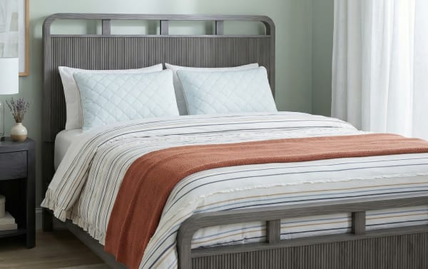 Queen-size bed with a gray wooden slatted headboard and footboard, white and beige striped bedding, light blue quilted pillows, and a rust-colored knitted throw blanket.