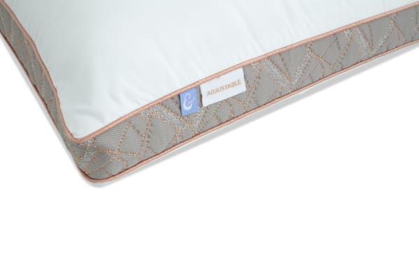 Adjustable pillow corner with white top, beige piping, and gray patterned side panel featuring fabric tags labeled "&" and "ADJUSTABLE."