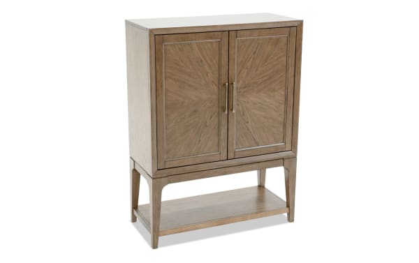 Wooden storage cabinet with two paneled doors featuring sunburst veneer pattern, brass handles, and lower open shelf, in light brown finish.