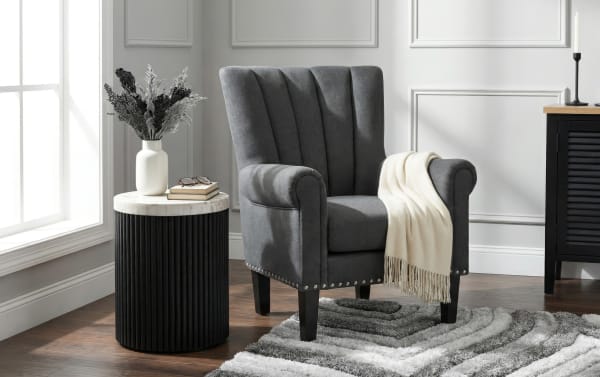 Gray upholstered accent chair with vertical channel tufting, nailhead trim, and a cream throw blanket, next to a black ribbed side table with a marble top and decorative vase.