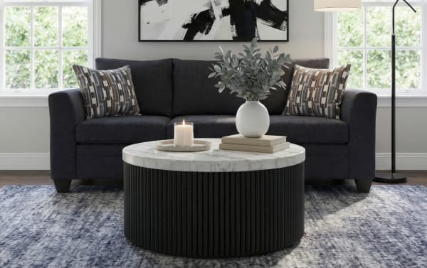Dark gray fabric sofa with patterned accent pillows, a round black ribbed coffee table with a white marble top, decorative vase with greenery, candle, and books in a modern living room.