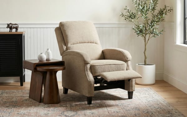 Beige fabric recliner chair with nailhead trim and dark wooden legs, shown with footrest extended, next to dark wood side tables and a potted plant in a bright living room.