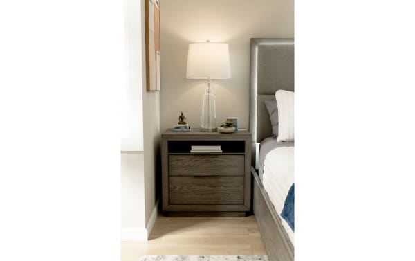 Modern gray wooden nightstand with two drawers, open shelf, and silver handles, topped with a clear glass table lamp and decorative items.