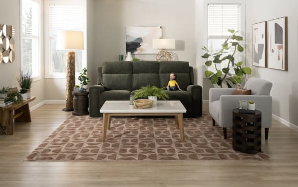 Modern living room with a dark green fabric sofa, light gray armchair, patterned beige rug, wooden coffee table with marble top, decorative plants, and abstract wall art.