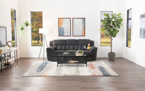 Modern living room with a dark gray fabric sofa, glass coffee table, abstract wall art, large indoor plant, and wooden flooring with a patterned area rug.