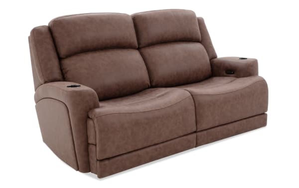 Brown leather reclining loveseat with cup holders on both armrests, cushioned backrests, and modern curved seat design.