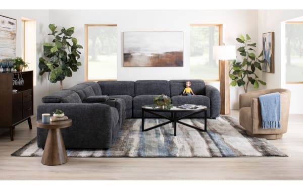 Modern living room with a dark gray fabric sectional sofa, round black metal coffee table, beige swivel chair with blue throw, wooden side table, and decorative plants.