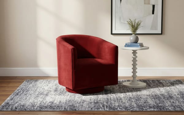 Red velvet swivel barrel chair with curved backrest on a gray patterned area rug, paired with a white round pedestal side table holding books and a vase.