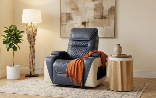 Modern blue and cream leather recliner chair with quilted stitching and cup holders, draped with an orange throw, next to a wooden side table and a floor lamp.