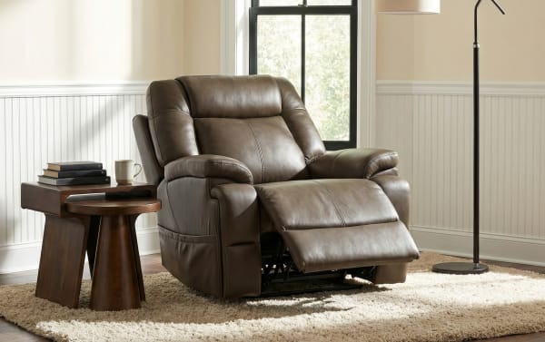 Brown leather recliner chair with padded armrests and cushioned backrest, paired with a wooden side table and floor lamp in a cozy living room setting.