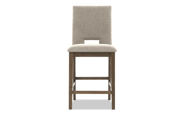 Upholstered beige fabric bar stool with cushioned seat, wooden frame, and rectangular cutout in backrest, modern dining chair design.