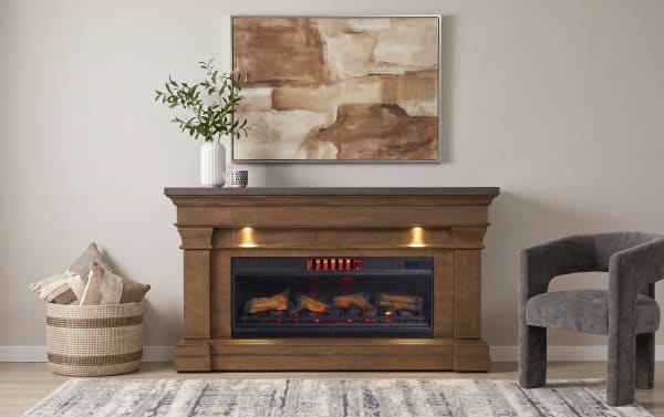 Modern electric fireplace with realistic glowing logs in a wooden mantel, beige walls, abstract brown artwork, woven basket with pillows, and gray upholstered chair.