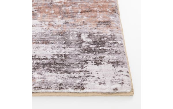 Close-up of a modern area rug corner with a low pile, featuring a distressed abstract pattern in muted gray, beige, and brown tones, with beige stitched edges.