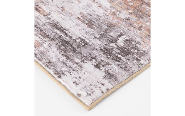 Close-up of a soft, textured area rug corner with a beige stitched edge, featuring a modern abstract pattern in shades of gray, white, and brown.