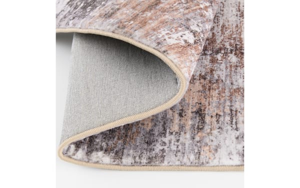 Close-up of a soft, modern area rug with a beige, brown, and gray abstract pattern, showing the textured fabric and stitched edges.