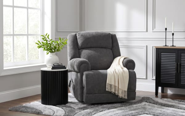 Gray fabric recliner chair with a cream throw blanket, black ribbed side table with white top, white vase with green leaves, and black cabinet in a modern living room.