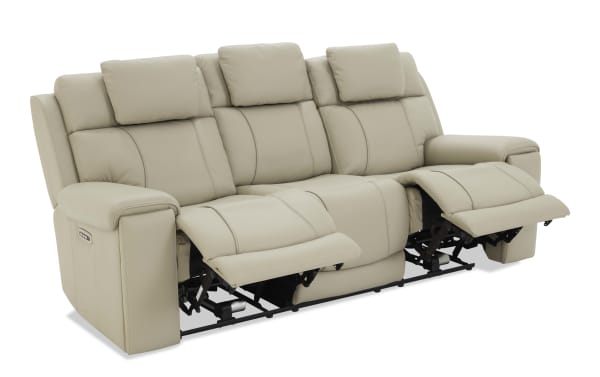 Beige leather reclining sofa with three seats, padded headrests, and armrests, featuring electric recline controls on the side.