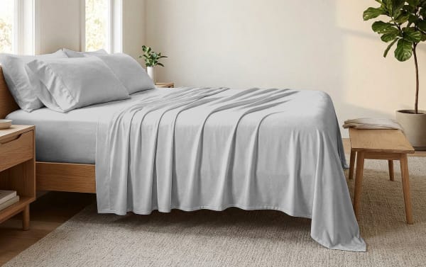 Modern wooden bed with light gray cotton sheets, matching pillowcases, and a soft blanket in a minimalist bedroom setting with natural light.