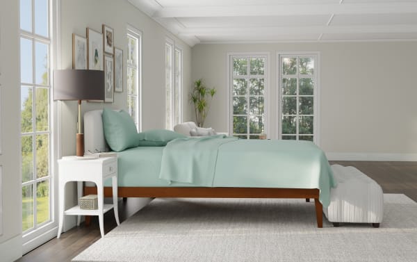 Modern bedroom with a wooden platform bed featuring light blue bedding, white nightstand with lamp, large windows, and a gray upholstered bench.
