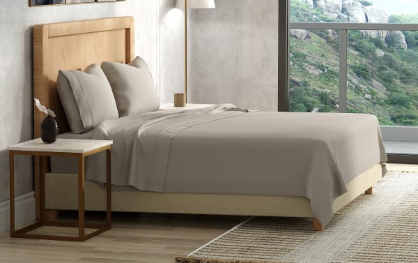Modern bed with beige fabric base, taupe cotton sheets, matching pillowcases, and a wooden headboard, next to a marble-top side table.
