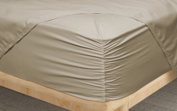 Beige fitted sheet with ruched corner detail on a light wood bed frame, showcasing smooth, wrinkle-resistant fabric ideal for bedroom decor.