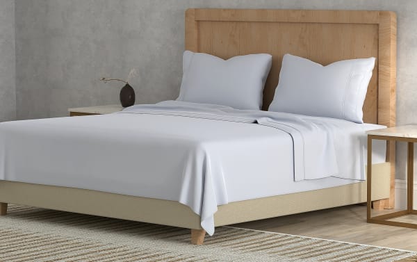 King-size bed with beige fabric base, light wood headboard, white cotton sheets, two white pillows, and modern side tables in a minimalist bedroom.
