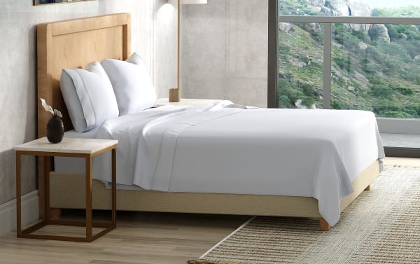 Modern bed with light wood headboard, beige upholstered base, white linens, two white pillows, and a marble-top side table with a small decorative vase.